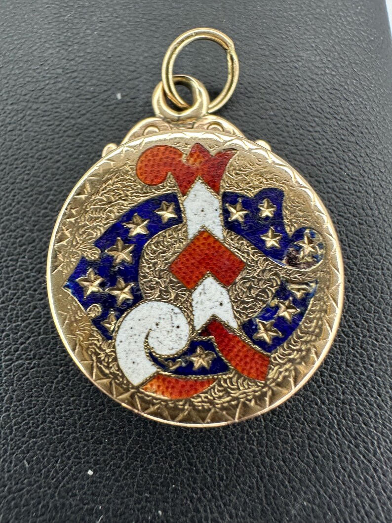 May include: A gold-toned pendant with a detailed design. The pendant features a central design in red, white, and blue, with a pattern of stars. The pendant has a loop at the top for attaching to a chain.