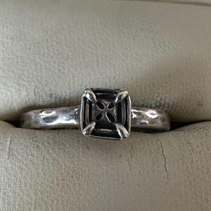 May include: A silver ring with a square, cross-shaped design. The ring has a textured band and a dark, detailed center. The ring is set against a neutral background.