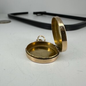 May include: A small, open, gold-colored locket with a hinged lid. The locket is round and has a simple design. The interior is also gold-colored. A small loop is attached for a chain.