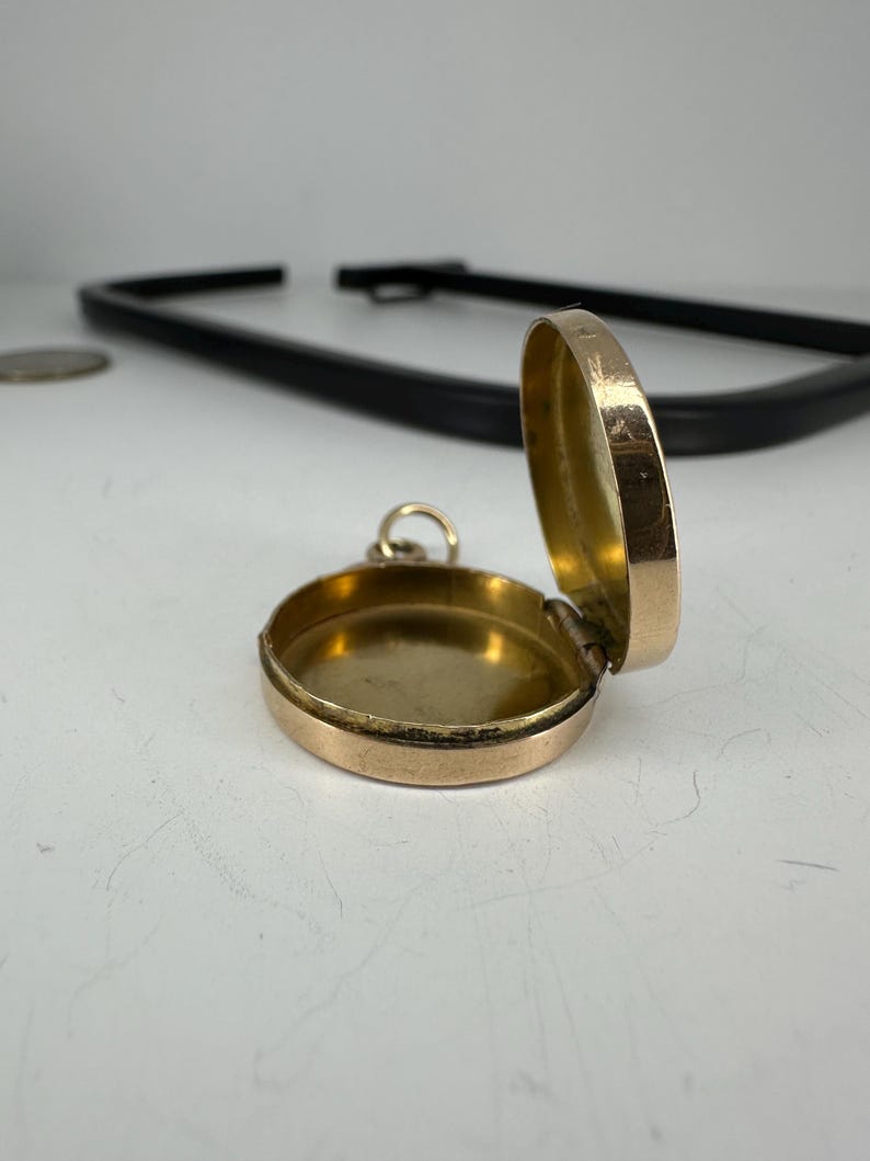 May include: A small, open, gold-colored locket with a hinged lid. The locket is round and has a simple design. The interior is also gold-colored. A small loop is attached for a chain.