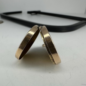 May include: Two gold-colored hoop earrings, slightly open, are the focal point of this image. The earrings have a smooth, rounded design. A black, curved object is in the background. The earrings are on a white surface.