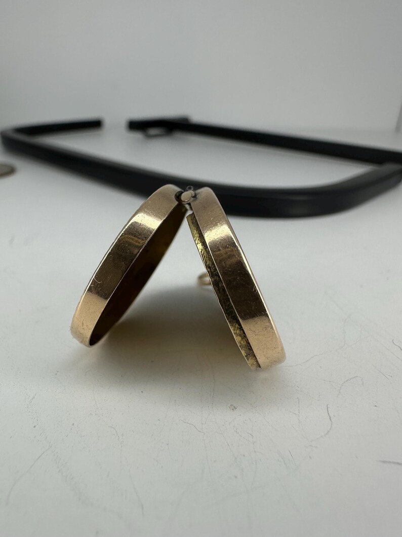 May include: Two gold-colored hoop earrings, slightly open, are the focal point of this image. The earrings have a smooth, rounded design. A black, curved object is in the background. The earrings are on a white surface.