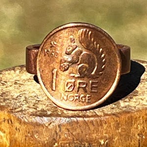 May include: A copper-colored ring with a 1 øre coin from Norway featuring a squirrel. The coin has the year 1950 and the text "1 øre NORGE" on it.