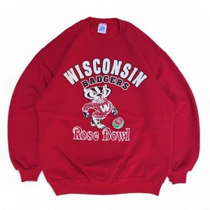 May include: Red sweatshirt featuring the text "WISCONSIN BADGERS" in white arched lettering above a cartoon badger mascot. The badger is wearing a red and white striped shirt and holding a rose. The text "Rose Bowl" is below the badger.