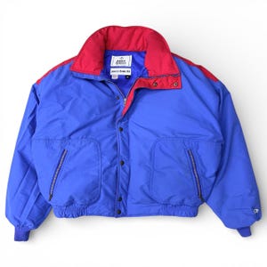 May include: A blue and red Alpine Designs goose down fill jacket. The jacket has a red collar and shoulder accents, with a blue body and two front pockets. The jacket has a zipper and snap closure. The brand name is visible on the collar.