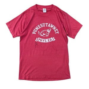 Puede incluir: Una camiseta roja con un gráfico blanco que presenta el texto "PUNXSUTAWNEY" encima de una ilustración de una marmota, y "PHYS. ED." debajo. La camiseta tiene cuello redondo y mangas cortas.