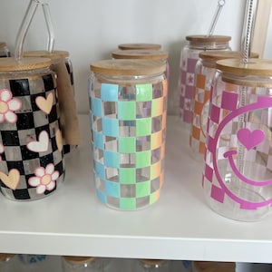 Puede incluir: Un estante con seis vasos de vidrio con tapas de bambú y pajitas reutilizables. Los vasos están decorados con diferentes patrones, incluyendo a cuadros, florales y caritas sonrientes.