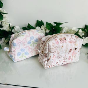 Puede incluir: Dos pequeños neceseres acolchados. Uno es blanco con lazos rosas y detalles florales azules. El otro es blanco con un estampado rosa con temática de ballet. Ambos tienen cremalleras doradas y se exhiben sobre una superficie blanca.