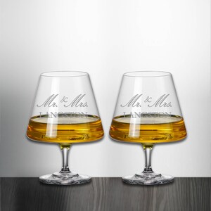 May include: Two clear glass whiskey glasses with a wide base and a flared top. The glasses are filled with amber liquid and have the text "Mr. & Mrs. LANGSTON" etched on the side.