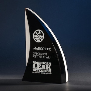 May include: Black and white crystal award with a shark fin shape. The award has the text "Marco Lex Specialist of the Year American Leak Detection The Original Leak Specialists" and a symbol of a water drop with a line through it.