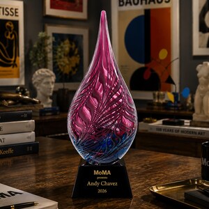 May include: A teardrop-shaped glass sculpture featuring a swirling pattern of magenta and blue hues. The sculpture is mounted on a black, square-based pedestal with the text "MoMA presents Andy Chavez 2026" printed in gold lettering on the front face.