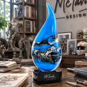 May include: A teardrop-shaped glass sculpture featuring a vibrant blue hue with swirling black and silver metallic accents. The sculpture is mounted on a black rectangular base with the white text McTavish DESIGNS printed on the front face.