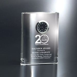 May include: A clear, rectangular award with a clock and the text "20 Years of Excellence." The award is presented to Monica Hadley and includes the AERON logo. The text also reads "Founder Award."