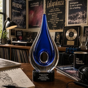 May include: A teardrop-shaped glass award featuring a deep blue center encased in clear glass, mounted on a black base. The base is engraved with the text: Producer of the Year, Presented to Donavan Marie, For Outstanding Contribution to Indie Music.
