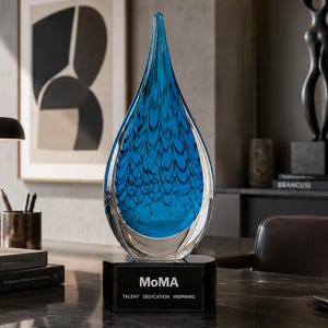 May include: A teardrop-shaped blue glass sculpture with a textured scale pattern inside, mounted on a black rectangular base. The base features the white text MoMA followed by the words TALENT DEDICATION INSPIRING printed on the front surface.
