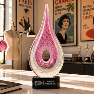 May include: A teardrop-shaped glass award featuring a clear outer layer and a textured pink core with an oval cutout. The award sits on a black rectangular base with the text 2026 FASHION WEEK PRESENTED TO MIRZA CHAVEZ printed in white.
