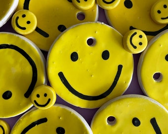 May include: A collection of yellow ceramic smiley face ornaments with black outlines. The ornaments have a hole in the center for hanging.