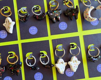 May include: A collection of hoop earrings displayed on black cards labeled hey jay studio. The earrings feature charms shaped like ghosts, cats, and crescent moons, decorated with colorful paint splatters in white, black, and metallic finishes.