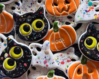 May include: A collection of ceramic Halloween ornaments featuring black cats with yellow eyes, orange pumpkins with carved faces, and white ghosts with colorful paint splatters. One orange pumpkin ornament has the word Boo! embossed on it.