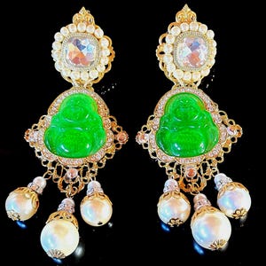 May include: A pair of ornate gold-toned earrings. Each earring features a green Buddha figure, surrounded by clear crystals and a gold filigree design. Large faux pearls dangle from the bottom, and a large crystal is at the top.