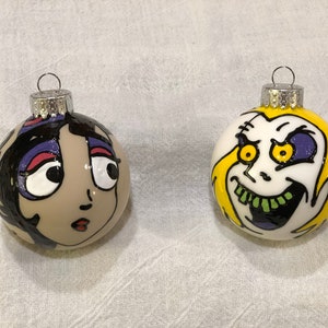 May include: Two hand-painted Christmas ornaments featuring cartoon faces. One ornament is yellow with a white face and green teeth. The other ornament is beige with a black haired woman's face.