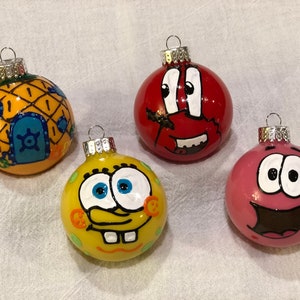 May include: Four hand-painted Christmas ornaments featuring characters from a popular animated series. The ornaments include a yellow pineapple house, a red crab, a yellow sponge, and a pink starfish. Each ornament has a silver cap.