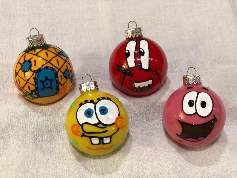 Op de afbeelding: Vier handgeschilderde kerstballen met personages uit een populaire animatieserie. De ballen omvatten een gele ananaswoning, een rode krab, een gele spons en een roze zeester. Elke bal heeft een zilveren dop.