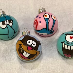 Op de afbeelding: Vier handgeschilderde kerstballen met stripfiguren. De ballen zijn rond en hebben zilveren metalen doppen. De karakters zijn onder meer een turquoise Squidward, een roze Gary de slak, een bruin karakter en een turquoise eenogig karakter.