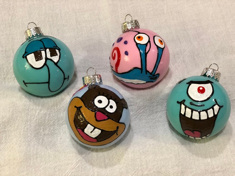 Op de afbeelding: Vier handgeschilderde kerstballen met stripfiguren. De ballen zijn rond en hebben zilveren metalen doppen. De karakters zijn onder meer een turquoise Squidward, een roze Gary de slak, een bruin karakter en een turquoise eenogig karakter.