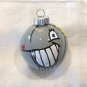 Op de afbeelding: Een grijze kerstbal met een cartoongezicht. De ornament heeft een grote witgetande grijns, blauwe ogen en een rood hart. Een zilveren metalen dop en ophanglus maken het ontwerp compleet.