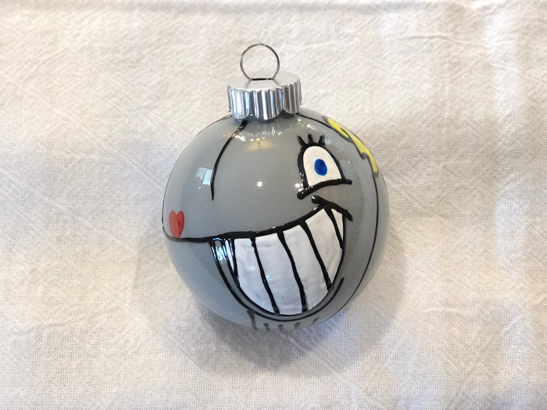 Op de afbeelding: Een grijze kerstbal met een cartoongezicht. De ornament heeft een grote witgetande grijns, blauwe ogen en een rood hart. Een zilveren metalen dop en ophanglus maken het ontwerp compleet.