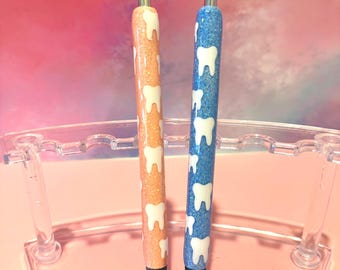 May include: Two glitter pens, one orange and one blue, each adorned with a pattern of white teeth. The pens are displayed in a clear plastic pen holder against a soft pink and purple background. The pens are designed for dental professionals or those in the dental field.