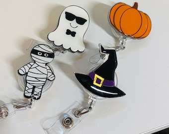 May include: Four Halloween-themed retractable badge holders. Designs include a white ghost with sunglasses, an orange pumpkin, a black witch hat with a purple band, and a mummy wrapped in white bandages. Each badge holder has a clear plastic strap and a metal clip.