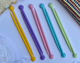 Può includere: Una collezione di cinque ferri da maglia colorati in giallo, viola, rosa, verde acqua e verde. Ogni ago ha un'estremità arrotondata e sfaccettata. Sono visibili anche filato bianco e un campione di maglia.
