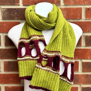 May include: A green knitted scarf with a ribbed texture. The scarf features burgundy and white patterned cutouts near the ends. The scarf is displayed on a white mannequin against a red brick wall.
