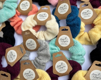 May include: A collection of knitted fabric hair scrunchies in various solid colors including yellow, black, pink, teal, cream, gray, and burgundy. Each scrunchie is attached to a brown cardboard tag featuring the text Loops & Layers.