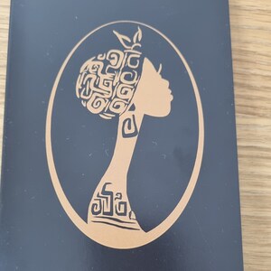 Könnte beinhalten: Eine schwarze Karte mit einer goldenen Silhouette einer Frau mit einem gemusterten Haarstil. Die Frau blickt nach rechts und hat ein gemustertes Design an Hals und Schulter.