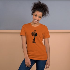 May include: A woman wearing an orange t-shirt with a black silhouette of a woman with an afro hairstyle.