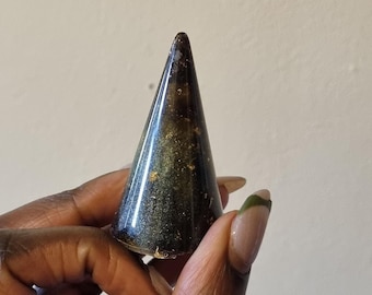 May include: A dark brown, cone-shaped object with gold flecks. The object is held in a hand and appears to be made of a polished, possibly natural material. The cone has a smooth, reflective surface.