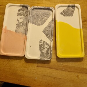 Op de afbeelding: Drie rechthoekige dienbladen van beton met een marmerpatroon. De dienbladen zijn in verschillende kleuren: roze, grijs en geel.