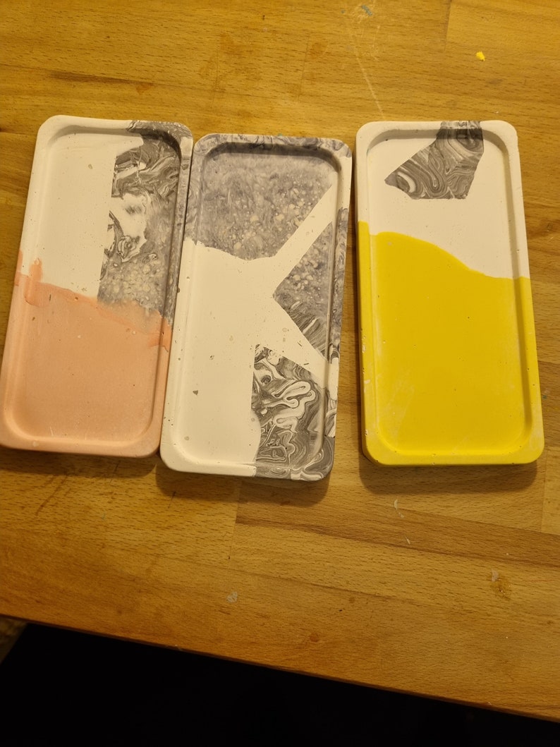Op de afbeelding: Drie rechthoekige dienbladen van beton met een marmerpatroon. De dienbladen zijn in verschillende kleuren: roze, grijs en geel.