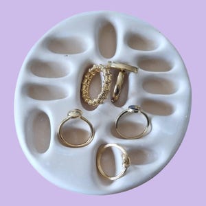Może przedstawiać: Biała ceramiczna podstawka na pierścionki z ośmioma indywidualnymi przegródkami do przechowywania pierścionków. Podstawka jest wypełniona złotymi pierścionkami, w tym obrączką, pierścionkiem z dużym kamieniem i pierścionkiem z mniejszym kamieniem.