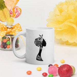 May include: White ceramic mug with a black silhouette of a woman with a patterned hairstyle. The woman is facing to the right and has a simple, elegant design.
