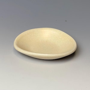 May include: A small, oval-shaped ceramic dish in a speckled, creamy beige color. The dish has a shallow, curved design and a smooth, rounded rim. The overall aesthetic is minimalist and organic.