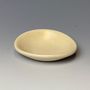May include: A small, oval-shaped, off-white ceramic dish. The dish has a slightly textured surface and a shallow, curved design. The color is a warm, creamy tone, and the dish appears to be handmade.