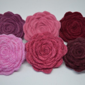 以下が含まれることがあります： ピンクとボルドーの色合いのフェルトの花の装飾が6つ。花は半円形に配置されており、一番濃い色が上、一番薄い色が下にあります。