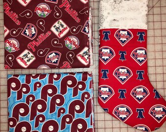 May include: Two pieces of fabric with a Phillies pattern, one red and white with the team logo and the other blue and maroon with the letter "P". A red and white stocking with the Phillies logo and the word "Phillies" is also shown.
