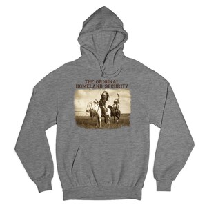 May include: A gray hooded sweatshirt with a sepia-toned graphic. The graphic depicts two Native Americans on horseback in a field, with the text "THE ORIGINAL HOMELAND SECURITY" above the image. The hoodie has a drawstring and a front pocket.
