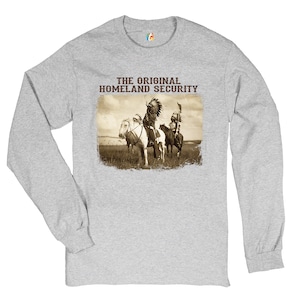 May include: A gray long-sleeve t-shirt with the text "THE ORIGINAL HOMELAND SECURITY" printed on the front. The text is in brown. The shirt features a vintage image of two Native American men on horseback.