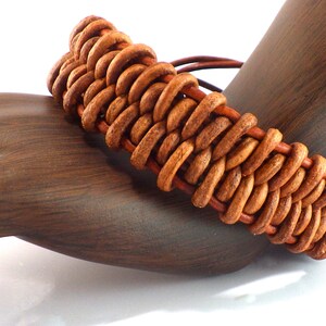 Puede incluir: Una pulsera de cuero marr&oacute;n hecha a mano con un dise&ntilde;o trenzado. La pulsera se exhibe en un soporte para joyas con forma de mano de madera, mostrando sus intrincados detalles y tonos c&aacute;lidos. El dise&ntilde;o de la pulsera presenta una serie de bucles de cuero interconectados.